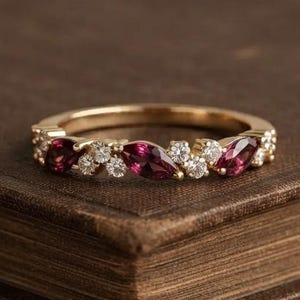 Può includere: Un anello in oro caratterizzato da una serie di pietre preziose rosso scuro taglio marquise alternate a piccole pietre rotonde trasparenti lungo la fascia. Il design presenta un motivo ripetitivo di pietre marquise rosse e pietre rotonde.