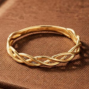 Op de afbeelding: Een dunne gouden ring met een delicaat gedraaid of gevlochten ontwerp. De band is gemaakt van gepolijst geelgoud, wat een doorlopend verweven patroon creëert dat om de vinger valt. De ring heeft een glad en reflecterend oppervlak.