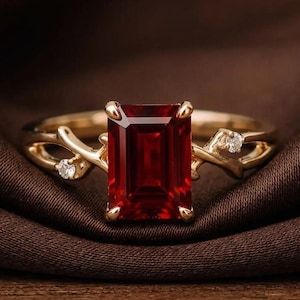 May include: A gold ring featuring a large, rectangular, emerald-cut deep red gemstone as the centerpiece. The band is crafted with a branch-like design, accented by two small, round, clear white stones set on either side of the main red gem.