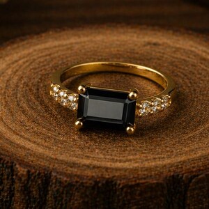 May include: A gold ring featuring a central rectangular emerald-cut black stone held by four prongs, with a band accented by small clear pavé stones on each side, showcasing a polished metallic finish and a refined, symmetrical design.