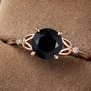 May include: A rose gold ring with a large, round black gemstone as the centerpiece. The band features delicate openwork designs and small accent stones on either side of the main stone. The ring is presented in a brown velvet ring box.