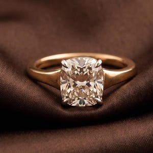 May include: A gold ring featuring a cushion-cut center stone held by four prongs. The band is smooth and polished, tapering slightly toward the setting. The stone is clear and faceted, reflecting light with a brilliant, multi-faceted appearance.