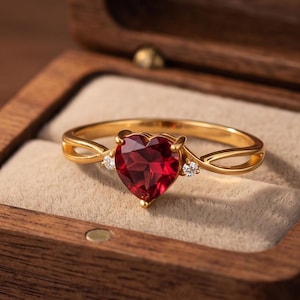 May include: A gold ring featuring a heart-shaped red gemstone at the center, flanked by two small clear stones on a split band. The ring is displayed inside a square wooden box with a beige velvet interior lining, showcasing a polished finish.