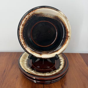 May include: A stack of three round ceramic plates featuring a dark brown center that transitions into a lighter cream and tan rim. The glaze has a glossy finish with a textured, mottled appearance along the edges of each plate in the set.