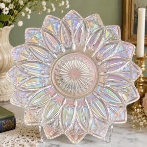 May include: A circular glass plate featuring an iridescent finish and a sunburst pattern. The design includes a central starburst motif surrounded by radiating petal-like shapes and diamond accents along the scalloped outer edge of the plate.