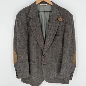 Puede incluir: Blazer de tweed marrón con coderas de ante y un alfiler de solapa a juego. La chaqueta tiene cierre de dos botones, dos bolsillos delanteros y botones en las mangas. El blazer está colgado de una percha de madera.