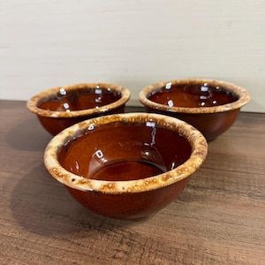 Op de afbeelding: Drie kleine, ronde keramische kommen met een glanzende donkerbruine binnenkant en een getextureerde, gespikkelde beige en bruine rand. De kommen hebben een gladde, reflecterende afwerking en een consistente vorm, gerangschikt in een driehoek.