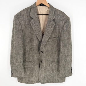 May include: A tweed blazer in shades of gray and brown hangs on a wooden hanger. The jacket has a notched lapel, two front pockets, and a two-button closure. The interior lining is a light tan color.