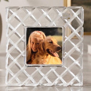 May include: A square Mikasa picture frame made of clear glass with a decorative lattice or woven ribbon pattern. The frame features a central square opening containing a photograph of a young child and a golden retriever dog. The glass has a smooth, reflective finish with frosted accents on the interwoven sections.