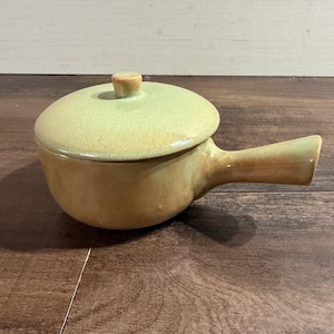 Può includere: Un pentolino in ceramica con coperchio, caratterizzato da una parte superiore smaltata in verde chiaro e una base color marrone chiaro. Il recipiente include un manico singolo che si estende lateralmente e un piccolo pomello centrato sul coperchio.