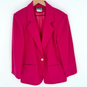 Puede incluir: Un blazer fucsia vibrante con un solo botón dorado. El blazer presenta una solapa con muescas clásica, dos bolsillos delanteros y el nombre de la marca "SAVANNAH" en la etiqueta. La chaqueta se muestra en una percha de madera.