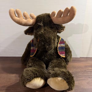 Pode incluir: Um peluche de alce com pelo castanho escuro, grandes hastes bege e patas bege. O alce usa um pequeno colete com um padrão de riscas multicoloridas com fios azuis, vermelhos e amarelos, rematado com um rebordo amarelo fino nas extremidades.