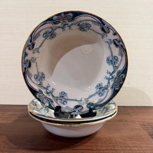 May include: A set of three stacked ceramic bowls featuring a white interior and a decorative rim pattern in dark blue and teal with gold accents. The bowls have a rounded shape and are displayed on a black stand, showcasing the intricate floral-inspired motifs along the edges of the top bowl.