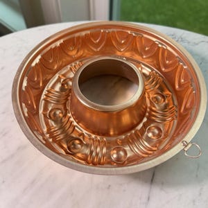 May include: A copper bundt pan with intricate embossed designs. The pan features a central tube and a decorative pattern around the edges. A small loop is attached to the side. The pan is made of copper and has a shiny finish.