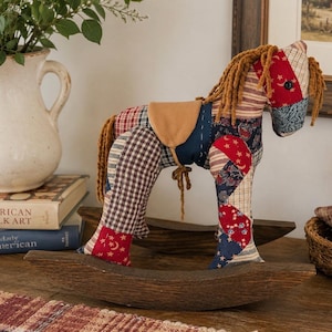 May include: A fabric rocking horse crafted from various patchwork patterns in red, white, and blue, featuring a tan saddle, dark brown wooden rockers, and a yarn mane and tail. The horse displays star, moon, and floral prints on its textile body.