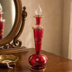 May include: A tall, deep red glass perfume bottle with a bulbous base and a slender neck. The bottle features delicate white floral patterns and gold painted accents. It is topped with a clear, pointed glass stopper with gold trim.