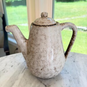 May include: A ceramic teapot with a glossy off-white glaze featuring a mottled brown speckled pattern. The teapot has a rounded body, a curved spout, a sturdy handle, and a matching lid with a small knob handle on top for easy removal.