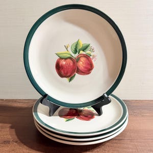 Puede incluir: Un juego de cuatro platos redondos de cerámica con centro color crema y borde verde oscuro. Cada plato presenta una ilustración central de dos manzanas rojas con hojas verdes y pequeñas flores blancas, representadas con un estilo realista.