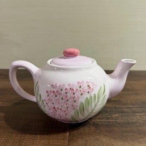 May include: A light pink ceramic teapot featuring a hand-painted floral design with clusters of small pink blossoms and elongated green leaves. The teapot includes a matching lid with a rounded pink knob and a curved handle and spout.