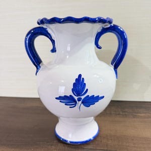 May include: A white ceramic vase featuring a glossy finish, two curved blue handles, and a scalloped rim. The body of the vase is decorated with a hand-painted blue floral motif consisting of three stylized leaves centered on the front surface.