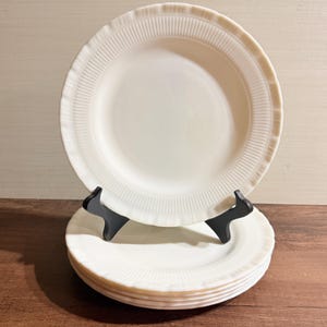 May include: A stack of four round, off-white milk glass plates. Each plate features a smooth center and a wide, raised rim decorated with a repeating vertical ribbed pattern and a scalloped edge. One plate is displayed upright on a black stand.