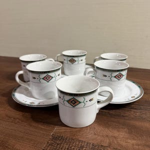 May include: A set of white ceramic mugs and matching saucers featuring a speckled finish and a decorative band with geometric patterns in green, orange, and black. Each mug has a curved handle and a cylindrical body, while the saucers have a matching border design.