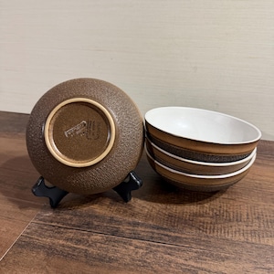Puede incluir: Un juego de cuatro cuencos redondos de cerámica con exterior marrón texturizado e interior blanco liso. Un cuenco se muestra sobre un soporte negro, revelando la base con el texto Denby Made in England. Los otros tres cuencos están apilados.
