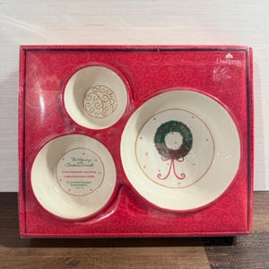 May include: A DaySpring boxed set containing three cream-colored ceramic bowls with red rims. One bowl features a green Christmas wreath, another has a gold swirl pattern, and the third displays the text The Meaning of the Christmas Wreath.
