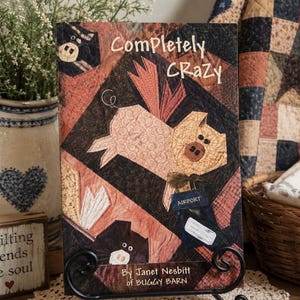 Può includere: Un libro intitolato Completely Crazy di Janet Nesbitt di Buggy Barn presenta una copertina in stile patchwork con l'illustrazione di un maiale volante. Il maiale è rosa chiaro con ali e indossa un'etichetta blu con la scritta AIRPORT su fondo marrone.