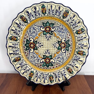 May include: A round ceramic plate with a scalloped edge, featuring a cream-colored base decorated with intricate hand-painted floral motifs in shades of dark blue, yellow, green, and red, arranged in a symmetrical pattern across the surface.
