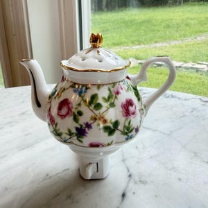 May include: White porcelain teapot night light with a floral rose pattern in pink, purple, and blue. The teapot has gold trim and a gold finial on the lid. The night light plugs directly into a wall outlet.