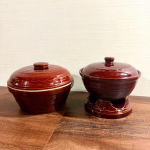 May include: Two dark brown glazed ceramic pots with matching lids. The left pot is a standard round bowl shape, while the right pot sits on a circular base with a cutout opening. Both feature embossed scalloped patterns along the rims and lids.