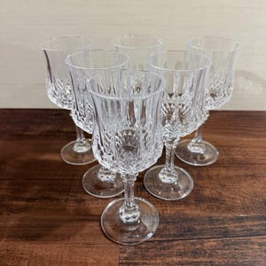 Può includere: Un set di sei calici in vetro trasparente caratterizzati da un motivo a taglio diamante sulla parte inferiore della coppa e da un bordo liscio e svasato. Ogni calice poggia su uno stelo sottile e sfaccettato collegato a una base circolare.