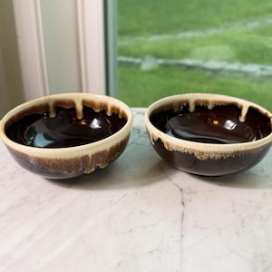 May include: Two ceramic bowls with a glossy brown interior and a cream-colored rim with a dripping effect. The bowls are placed side by side on a marble surface. The bowls are likely for serving food.