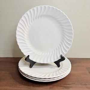 Puede incluir: Un juego de tres platos de cerámica blanca con acabado brillante y un patrón acanalado en espiral a lo largo del borde exterior. Los platos están apilados y el superior se mantiene en posición vertical mediante un soporte de plástico negro.