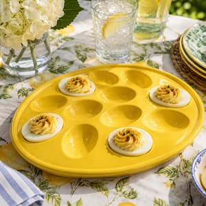 May include: A bright yellow ceramic deviled egg serving platter with twelve oval-shaped indentations. Four of the indentations are filled with halved hard-boiled eggs topped with piped yellow yolk filling and a sprinkle of green herbs.