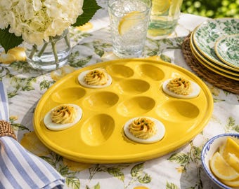 May include: A bright yellow ceramic deviled egg serving platter with twelve oval-shaped indentations. Four of the indentations are filled with halved hard-boiled eggs topped with piped yellow yolk filling and a sprinkle of green herbs.