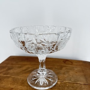 May include: A clear, stemmed glass dessert bowl with a detailed, etched floral pattern. The bowl has a wide, scalloped rim and sits on a pedestal base. The glass is transparent, allowing light to pass through, and is set on a wooden surface.