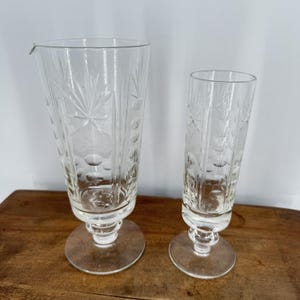 May include: Two clear, etched glass cocktail glasses. The taller glass has a spout. Both glasses have a decorative pattern of starbursts, circles, and lines. The glasses sit on a round base and are displayed on a wooden surface.