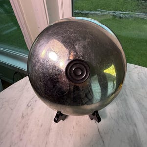 Puede incluir: Una tapa circular de metal plateado pulido con una superficie lisa y reflectante y un pomo negro circular en el centro. La tapa se apoya sobre dos pequeños pies negros en ángulo en la base, lo que le permite mantenerse en posición vertical.