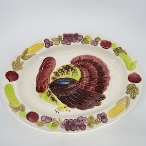May include: Oval ceramic serving platter with a hand-painted turkey design. The turkey is brown, red, and green, with a blue eye. The platter is decorated with corn, grapes, and other vegetables around the rim.