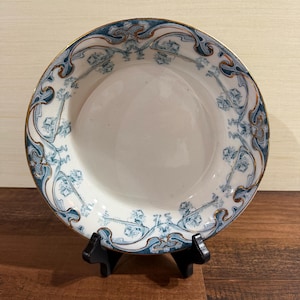 May include: A round ceramic bowl with a white center and a wide rim featuring a blue and white floral pattern with gold-toned trim. The bowl is displayed on a black plastic stand, showcasing the intricate decorative border and smooth finish.