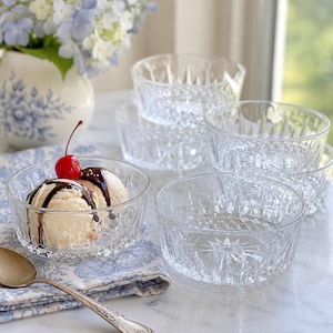 May include: A set of clear glass bowls featuring a faceted, starburst-patterned base and vertical ribbing along the sides. One bowl contains two scoops of vanilla ice cream topped with chocolate syrup and a single red maraschino cherry.