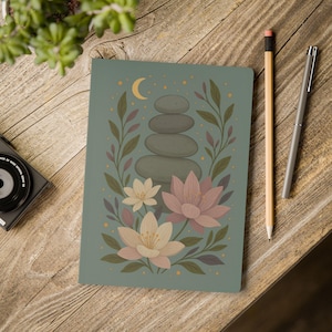 Può includere: Un diario turchese con un design floreale e di pietre. La copertina presenta una pila di pietre grigie, fiori di loto e una luna crescente. Una matita e una penna poggiano su una superficie di legno.