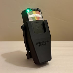 May include: A black paranormal activity detector with a green light illuminated. The device has a clip and the words "PARANORMAL SUPPLY" printed on the front. A label displays a color-coded scale and the text "mGAUSS(mG)".