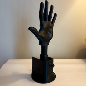 May include: A matte black 3D printed hand sculpture mounted on a cylindrical post attached to a curved base. The hand is positioned with fingers extended upward, and the base features a small circular knob on the side for adjustment.