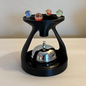 May include: A black, 3D-printed marble holder with a silver bell at the base. The holder has a unique, curved design and holds four colorful marbles. The marbles are blue, orange, red, and green. The bell has a small, decorative top.