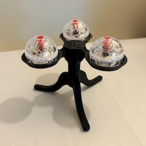 May include: A black, three-legged decorative stand with three clear, spherical lights. Each light has a red center and a faceted, clear cover. The stand is made of a matte black material and is on a white surface.