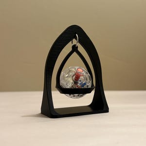 May include: A black, arched, decorative stand holding a clear, spherical object. The sphere has a red and silver interior. The stand is made of a solid material and has a flat base. The overall design is modern and minimalist.