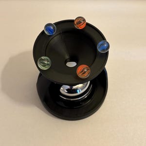 May include: A black marble run toy with six colorful marbles. The marbles are blue, orange, and green with swirling patterns. The toy has a funnel-shaped top and a circular base.